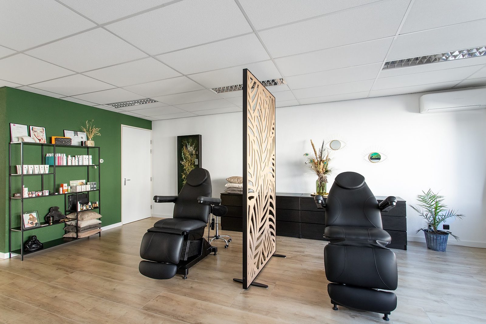 De salon: groene wand, twee zwarte behandelstoelen en een houten scheidingswand met bladmotief